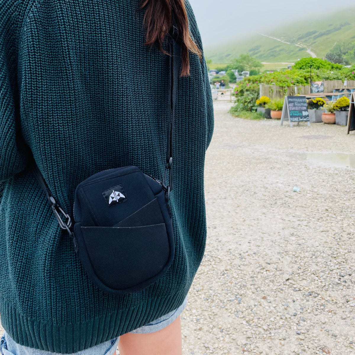 Recycled crossbody bag with adjustable strap made recycled wetsuits worn in a park seaside setting.