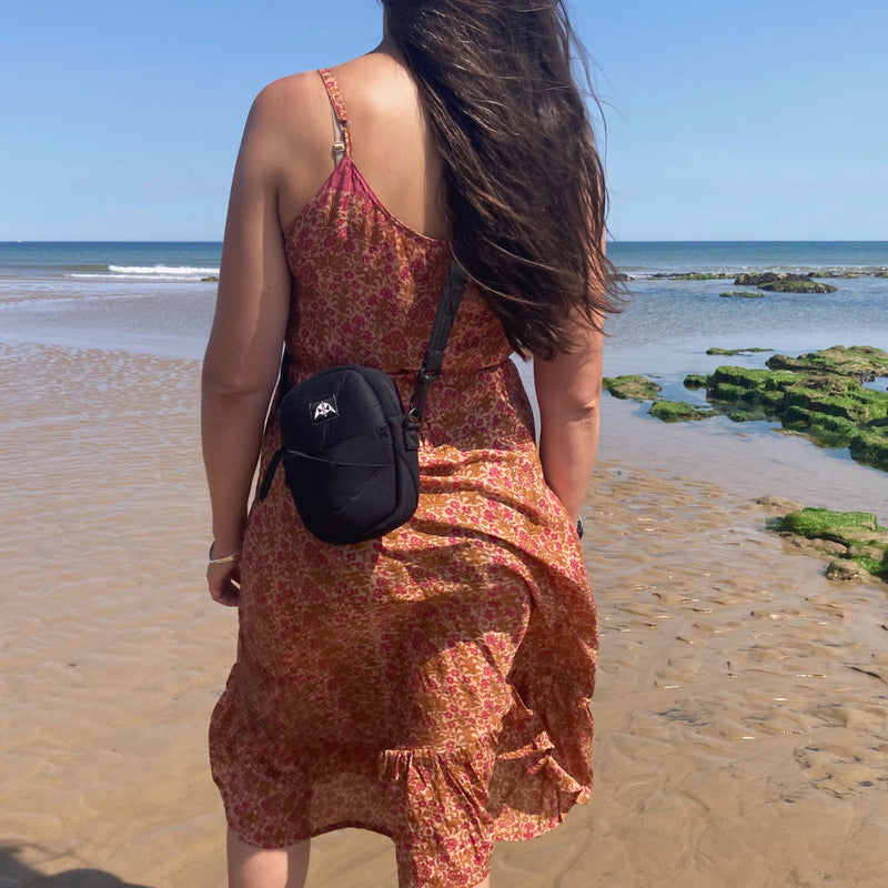 Recycled crossbody bag with adjustable strap and zipped closure worn on a beachside setting. 
