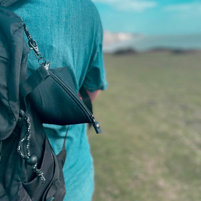 Person wearing a teal shirt with a pyramid shaped pouch (made from upcycled wetsuits)  over their shoulder, standing in a grassy field.