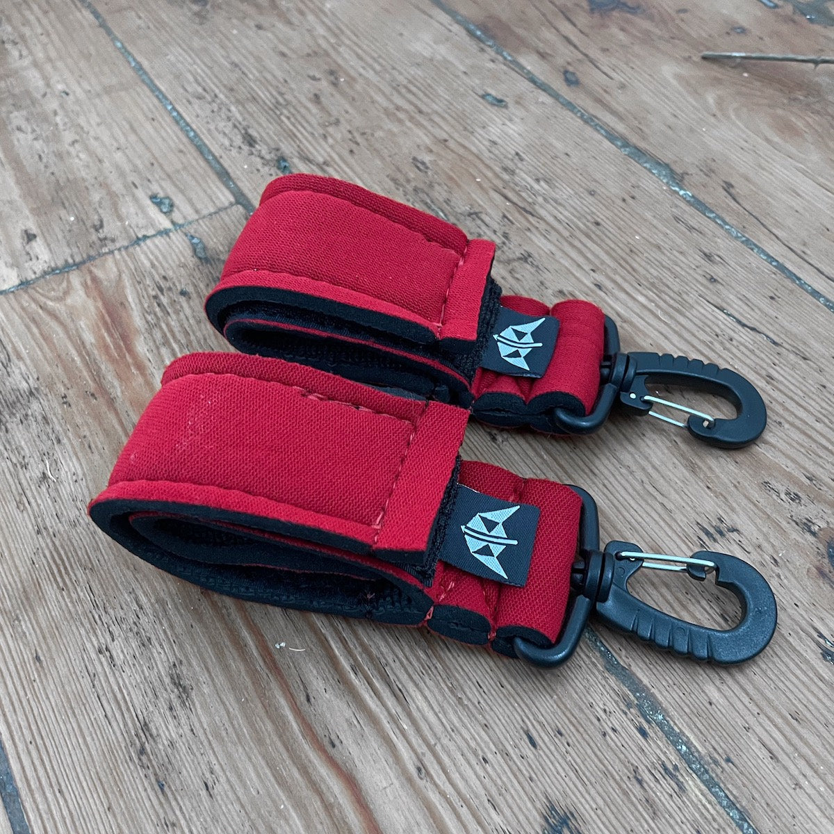A pair of red velcro SUP. paddle keepers with clips, placed on a wooden surface. Made from recycled wetsuits.