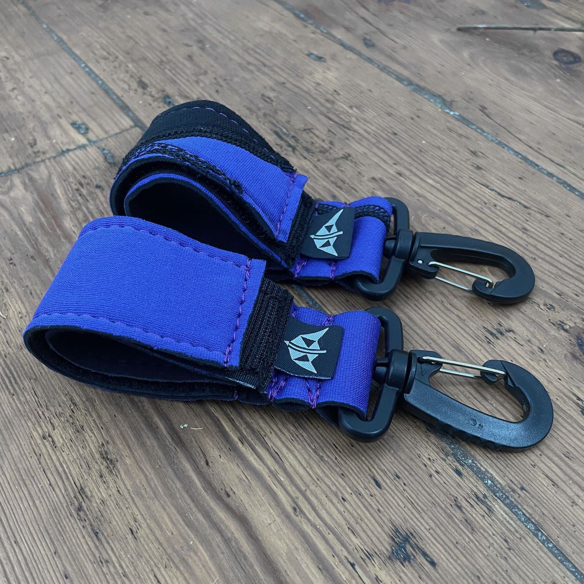 A pair of blue and black velcro paddle keepers with clips (recycled from wetsuits), placed on a wooden surface.
